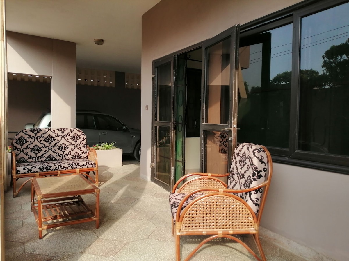 An outdoor seating area is featured with two patterned armchairs and a glass-topped coffee table. The space is bordered by large windows allowing natural light, and a vehicle is visible in the background, emphasizing accessibility.