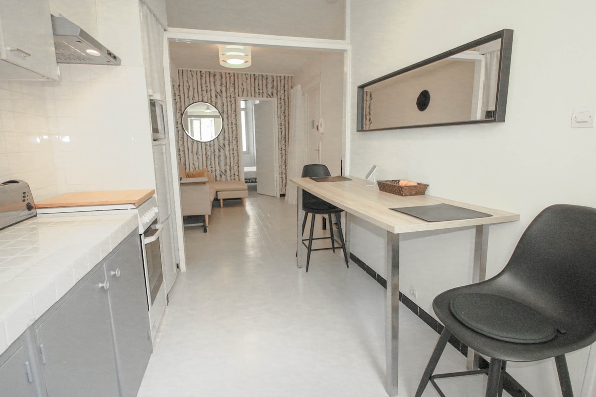 A modern kitchen space features sleek cabinetry and tiled countertops. A kitchen island with stools provides seating, while an oven and a toaster are visible. The layout opens to a bright hallway, providing a glimpse of additional living areas in the distance.