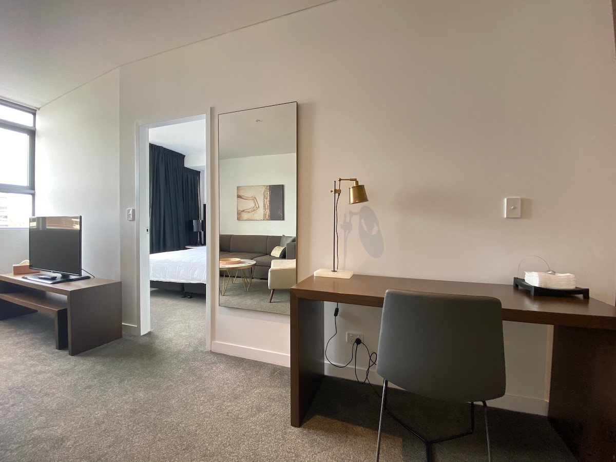 A modern room features a large mirror reflecting a seating area in the background. A desk is positioned against the wall, complemented by a stylish lamp. A flat-screen TV is placed on a low cabinet, contributing to the clean lines of the space.