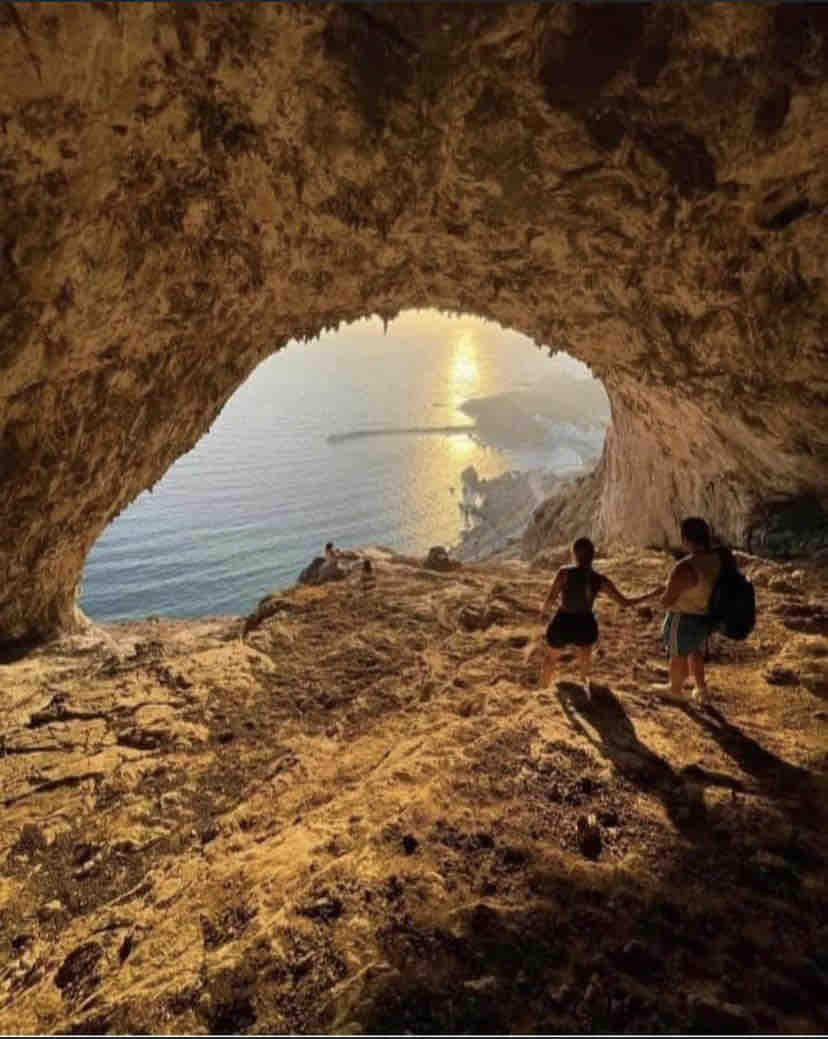 A cave entrance frames a serene view of the sea and distant shoreline, bathed in warm sunlight. Two figures stand near the cave's opening, their silhouettes contrasting against the illuminated ocean. The rocky terrain inside the cave features earthy tones, enhancing the natural setting.