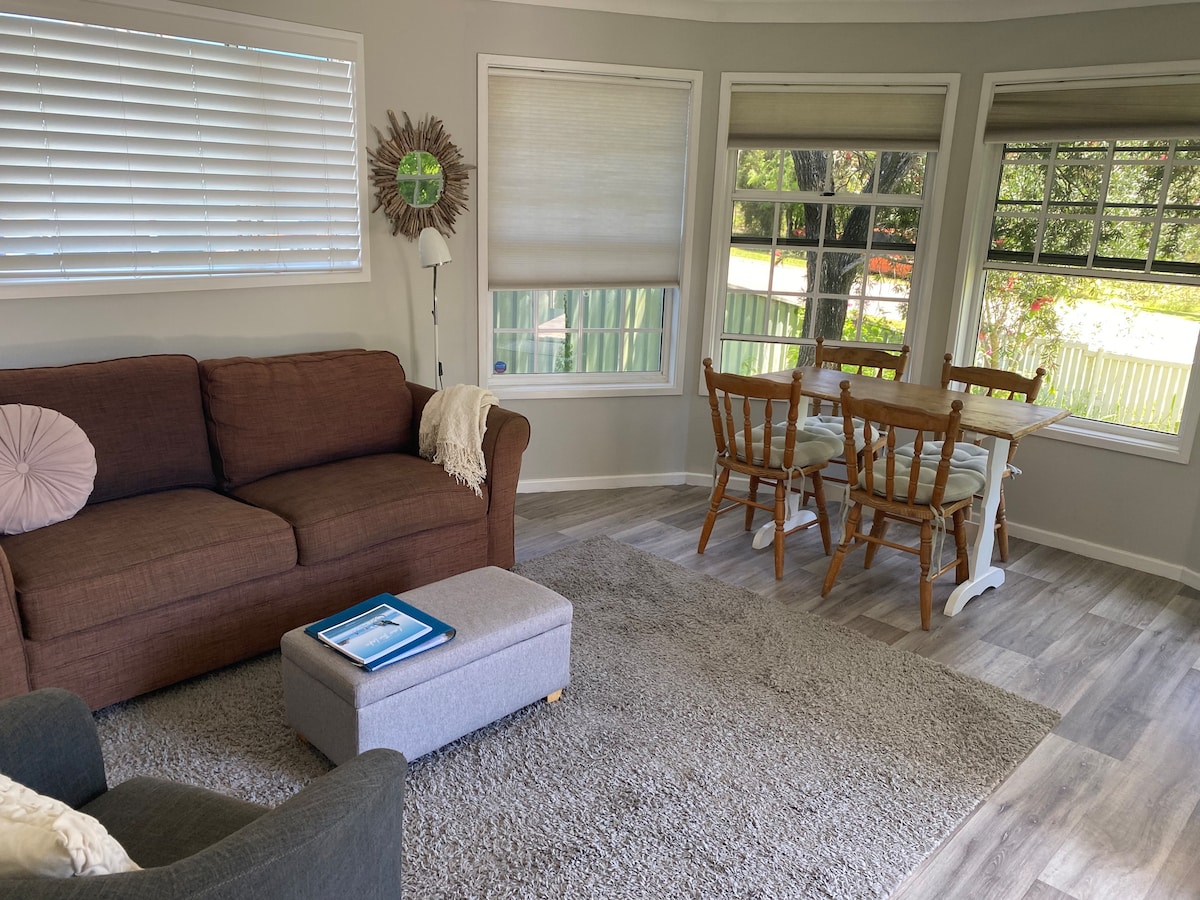 A light-filled lounge area features a brown sofa and a cozy armchair, accompanied by an ottoman. A small dining table with wooden chairs is positioned near large windows, inviting natural light into the space. Neutral-colored walls and carpet create a calming environment.