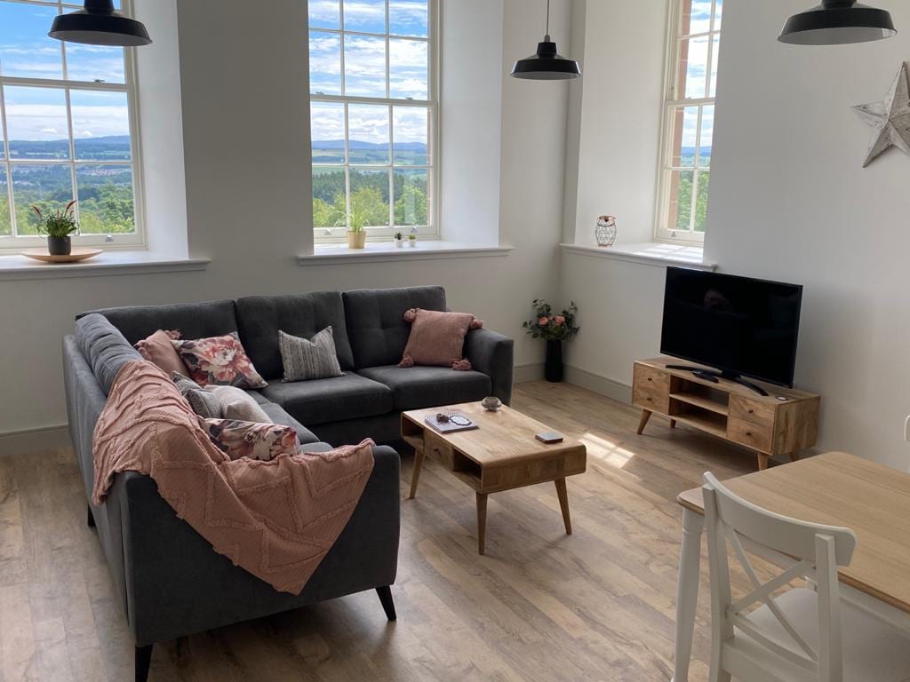 A spacious living area is illuminated by natural light from large windows, offering views of the surrounding landscape. A gray sectional sofa, adorned with cushions and a throw blanket, faces a wooden coffee table and a television stand. A dining table is positioned nearby.