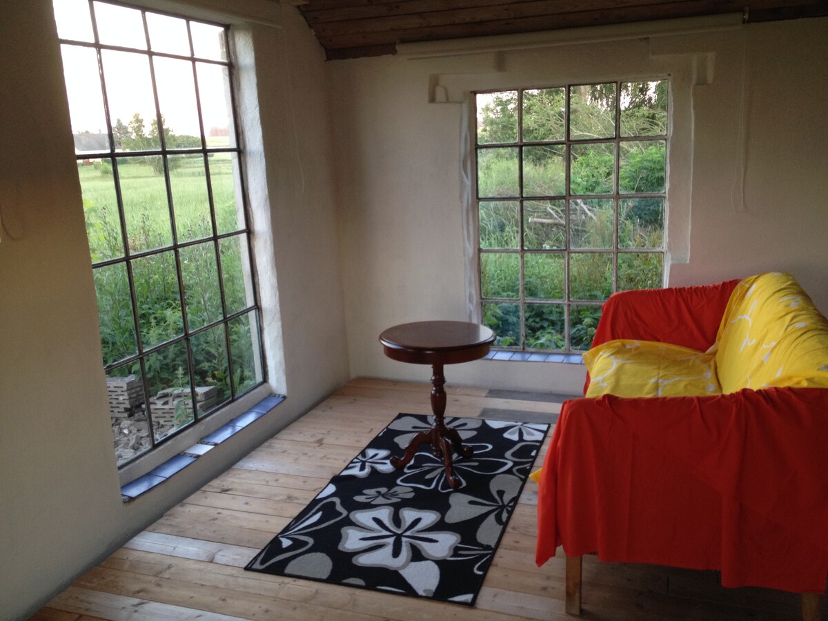 A comfortable seating area features a bright couch draped with a red throw and a small circular table. Large windows allow for ample natural light and offer views of the surrounding greenery. A patterned rug enhances the inviting atmosphere of the space.