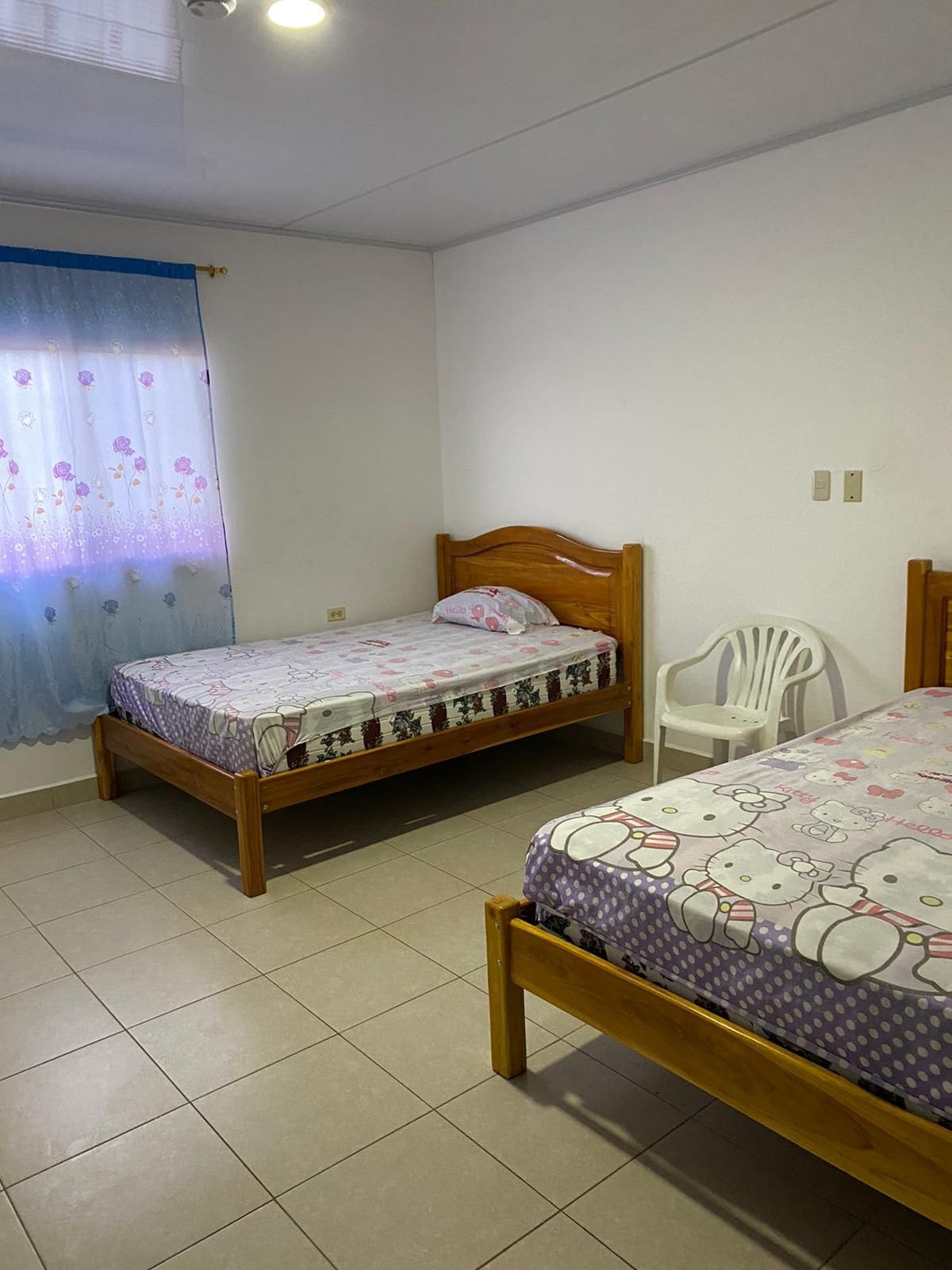 A bright bedroom displays two wooden beds with colorful bedding. A plastic chair is positioned nearby, and a light blue curtain softly filters sunlight through a window. The tiled floor adds to the clean and simple ambiance of the room.