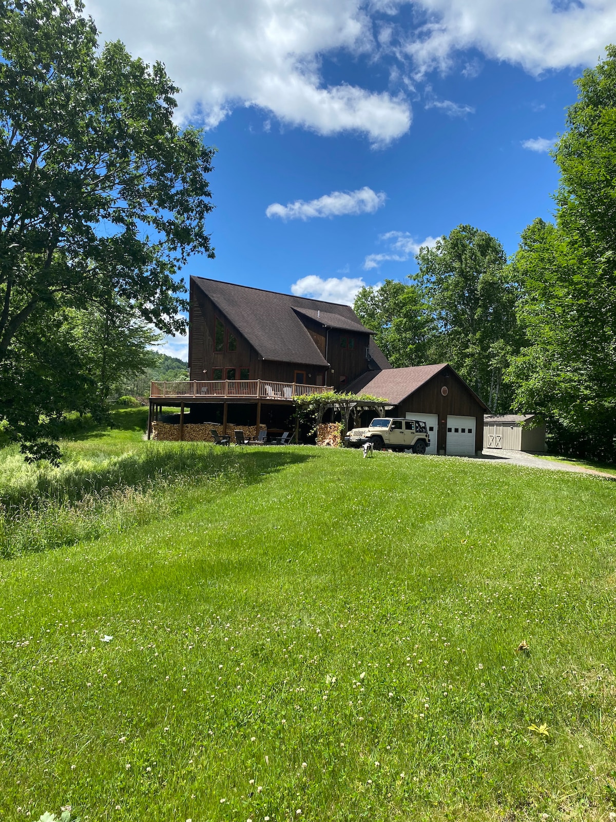 A multi-story house is set on a spacious green lawn, surrounded by trees. The structure features a large deck and a fully accessible driveway, accommodating multiple vehicles. A clear blue sky enhances the scenery, creating a calm and open atmosphere.
