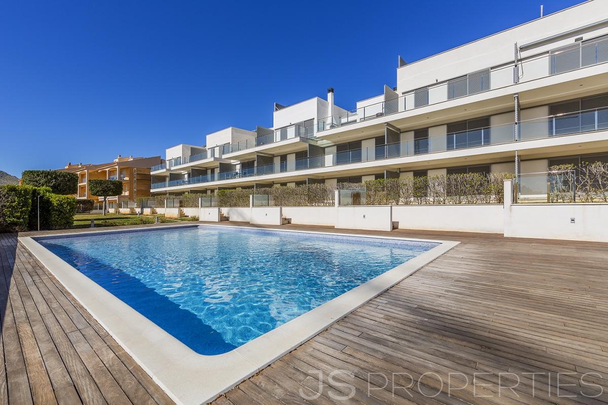 A clear blue swimming pool is set within a spacious wooden deck, surrounded by well-maintained gardens. The modern apartment complex is visible in the background, featuring multiple balconies that overlook the pool area. The bright sky enhances the inviting outdoor environment.
