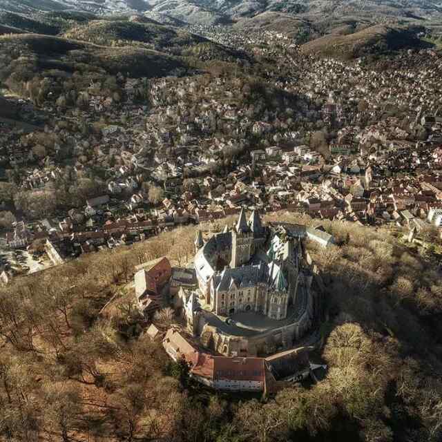 An aerial view showcases a medieval castle surrounded by lush greenery and rolling hills. The castle features distinctive towers and turrets, while nearby, a small town with clustered rooftops is visible, nestled among the trees and hills, enhancing the timeless charm of the landscape.