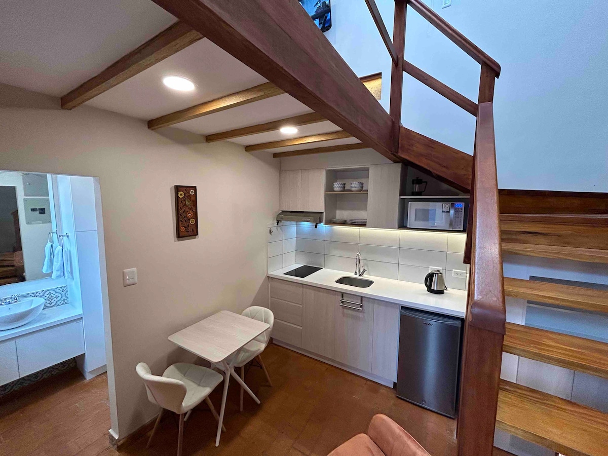 The kitchenette is equipped with modern appliances and minimalistic cabinetry. A small dining table with two chairs is positioned beside the kitchen. Warm wood accents line the ceiling, while a staircase leads to the lofted area. A sleek, shared bathroom is visible through an open doorway.