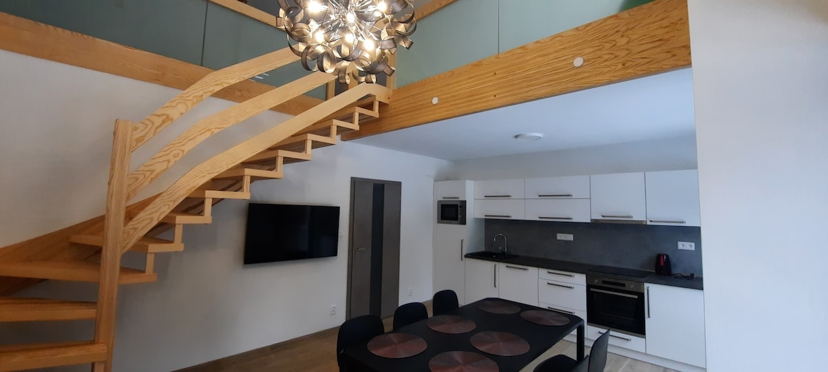 A modern, open-plan living area is featured, showcasing a wooden staircase leading to an upper level. A large dining table is surrounded by black chairs, while a sleek kitchen with white cabinetry is visible in the background. A flat-screen TV is mounted on the wall.