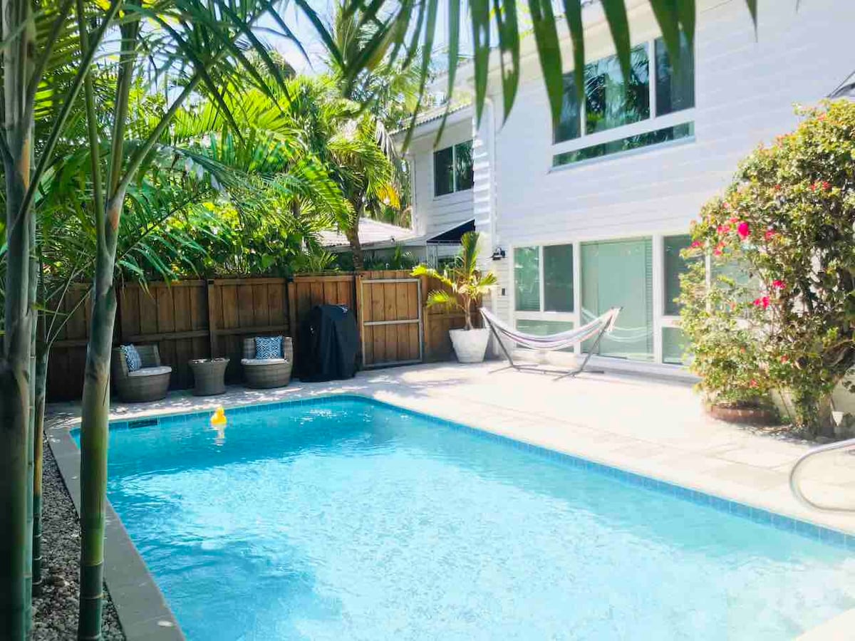 A serene outdoor space features a swimming pool surrounded by lush palm trees and vibrant plants. A hammock is positioned nearby, and a barbecue grill can be seen under the covered area. Natural light reflects off the water, creating a calming atmosphere.