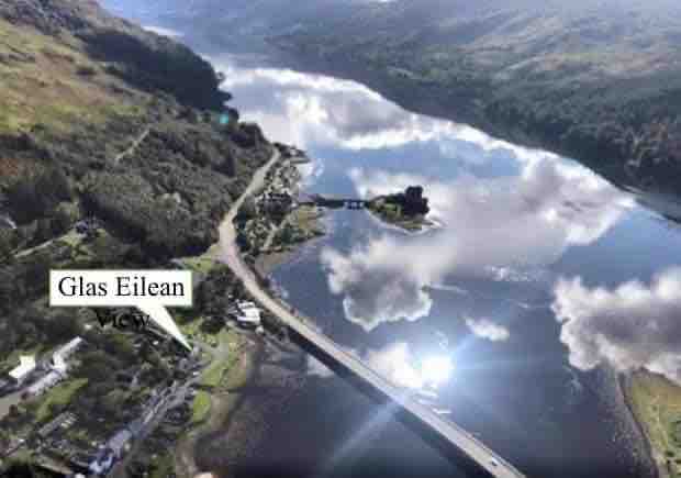 An aerial view captures the scenic surroundings of Glas Eilean, highlighting the tranquil waters of Loch Long and the nearby Eilean Donan Castle. The lush hills frame the landscape, while the village is nestled along the shore, showcasing the beauty of this lochside location.