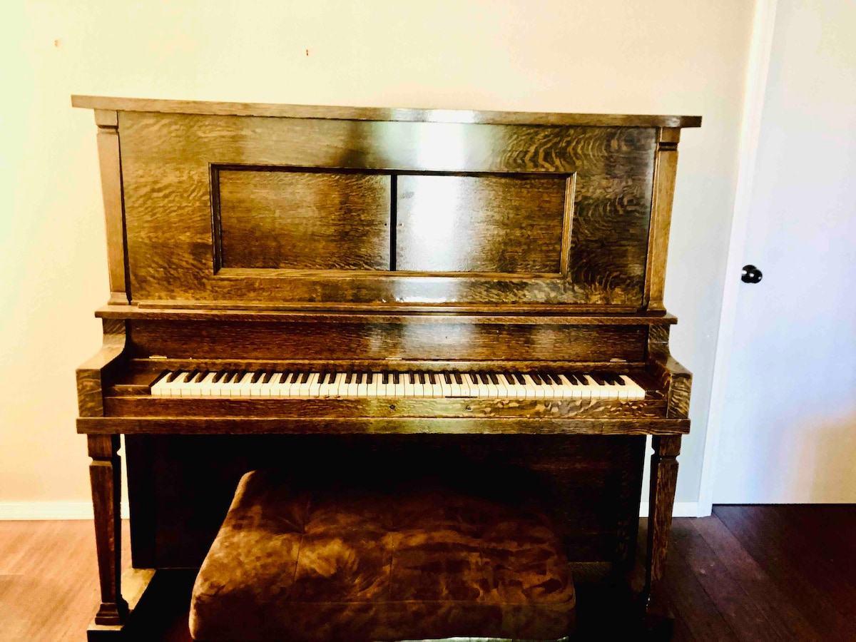 A classic wooden piano with a polished finish is positioned against a plain wall. The keys are visible, showcasing their ivory and black contrast. A cushioned bench sits in front of the piano, offering a comfortable place for playing.