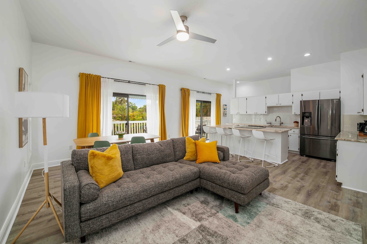 The living area features a spacious sectional sofa adorned with yellow accent pillows. Light wood floors complement the bright decor, which includes large windows with sheer curtains. A modern dining area with a table and chairs is visible, along with a well-equipped kitchen showcasing stainless steel appliances.
