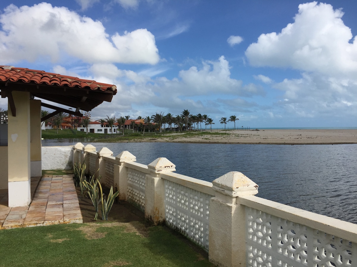 A side view of the house reveals a terracotta-tiled roof and a lush green lawn leading to a decorative white wall. Beyond the wall, a peaceful lagoon reflects the clear blue sky and palm trees, while a sandy beach stretches along the horizon.