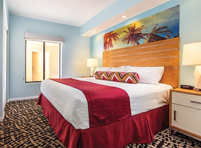 A spacious sleeping area features a large bed with crisp white bedding and a decorative red throw. A vibrant mural depicting palm trees adorns the wall above the headboard. Soft light from two bedside lamps enhances the relaxed atmosphere, while a large window provides natural light.