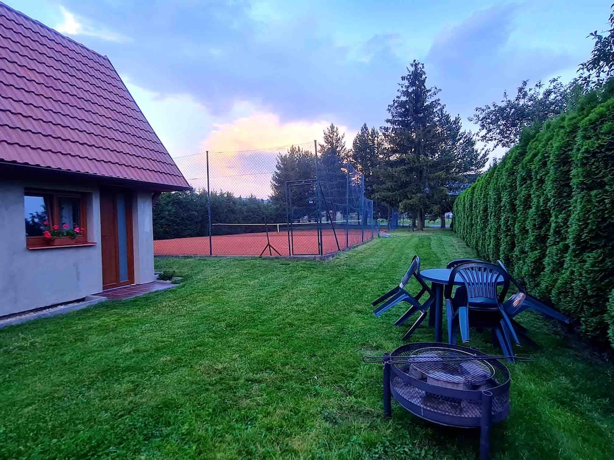 A well-maintained outdoor space features a circular fire pit surrounded by seating. A dining table and chairs are positioned on the lawn, which adjoins a tennis court, partially shaded by tall hedges. The sky is painted in soft colors at dusk.