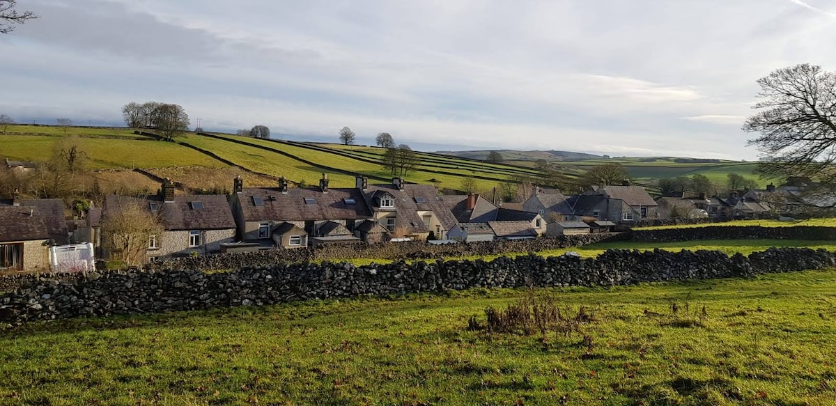 A scenic view of a village nestled among rolling green hills. Traditional stone houses are aligned along a winding road, with dry stone walls serving as boundaries for fields. Trees dot the landscape, and the sky is lightly clouded, enhancing the tranquil atmosphere.