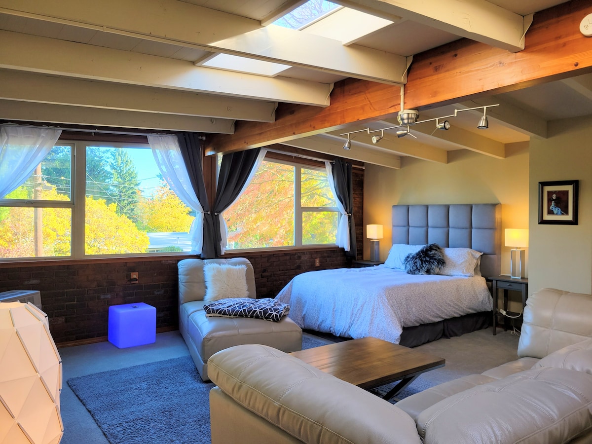 A spacious loft features large windows allowing natural light to fill the room. A comfortable bed with a tufted headboard is complemented by bedside tables with lamps. A plush couch and a modern coffee table are arranged on a soft rug, creating a cozy lounge area.