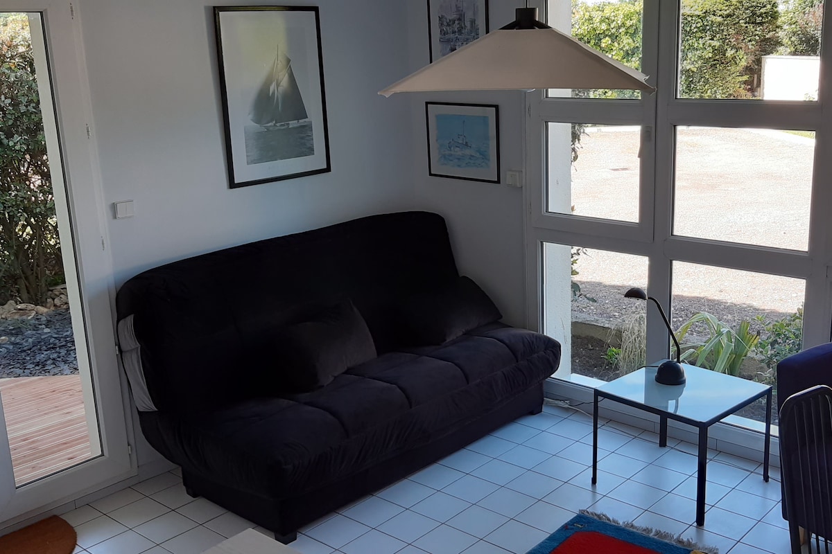 A cozy seating area features a dark sofa bed positioned near large windows, allowing ample natural light. A small table stands beside the sofa, accompanied by a modern lamp. Framed artwork adorns the walls, enhancing the inviting environment.