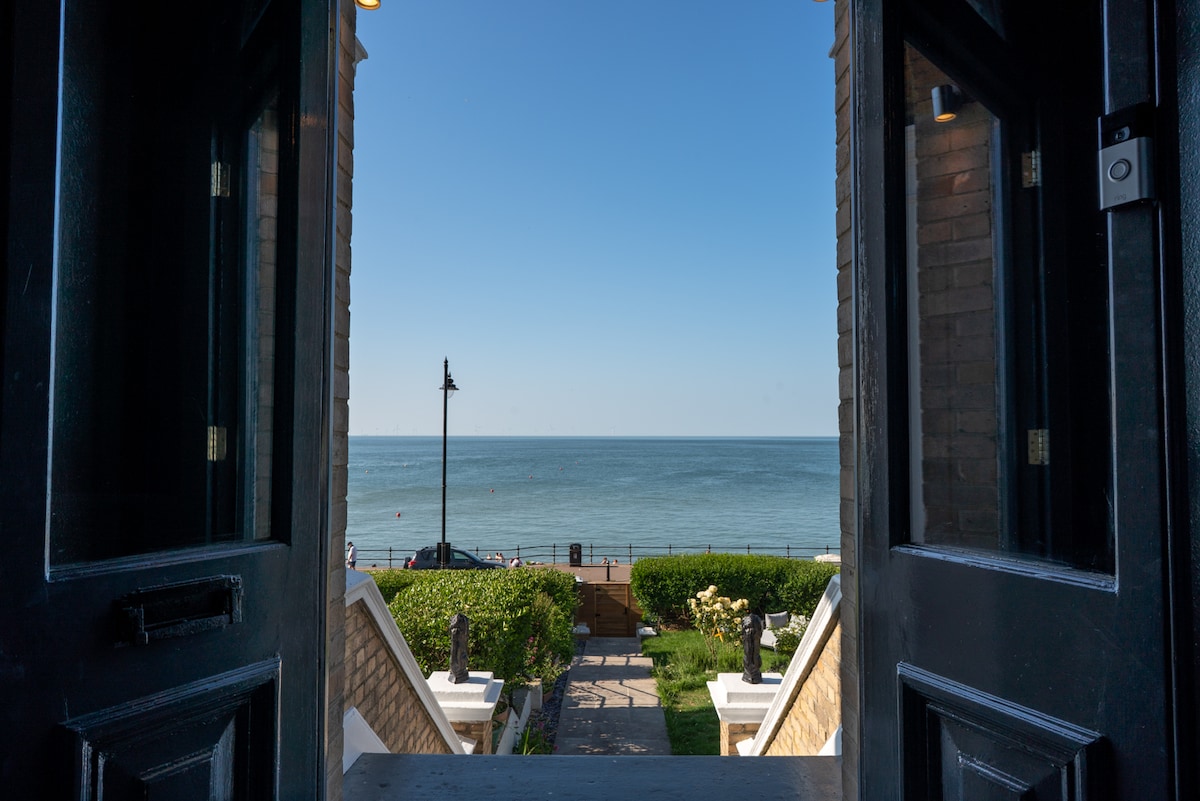 The view is framed by the open double doors, leading to a clear blue sky and the calm sea beyond. A pathway lined with greenery is visible, leading down to the beachfront, capturing a serene coastal scene.