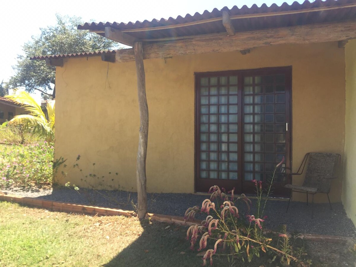 A rustic exterior of a single-story building is displayed, featuring natural earth-toned walls and wooden elements. Large, grid-patterned glass doors invite light, while a simple chair is positioned nearby. Lush greenery and blooming plants surround the structure, contributing to a serene outdoor setting.