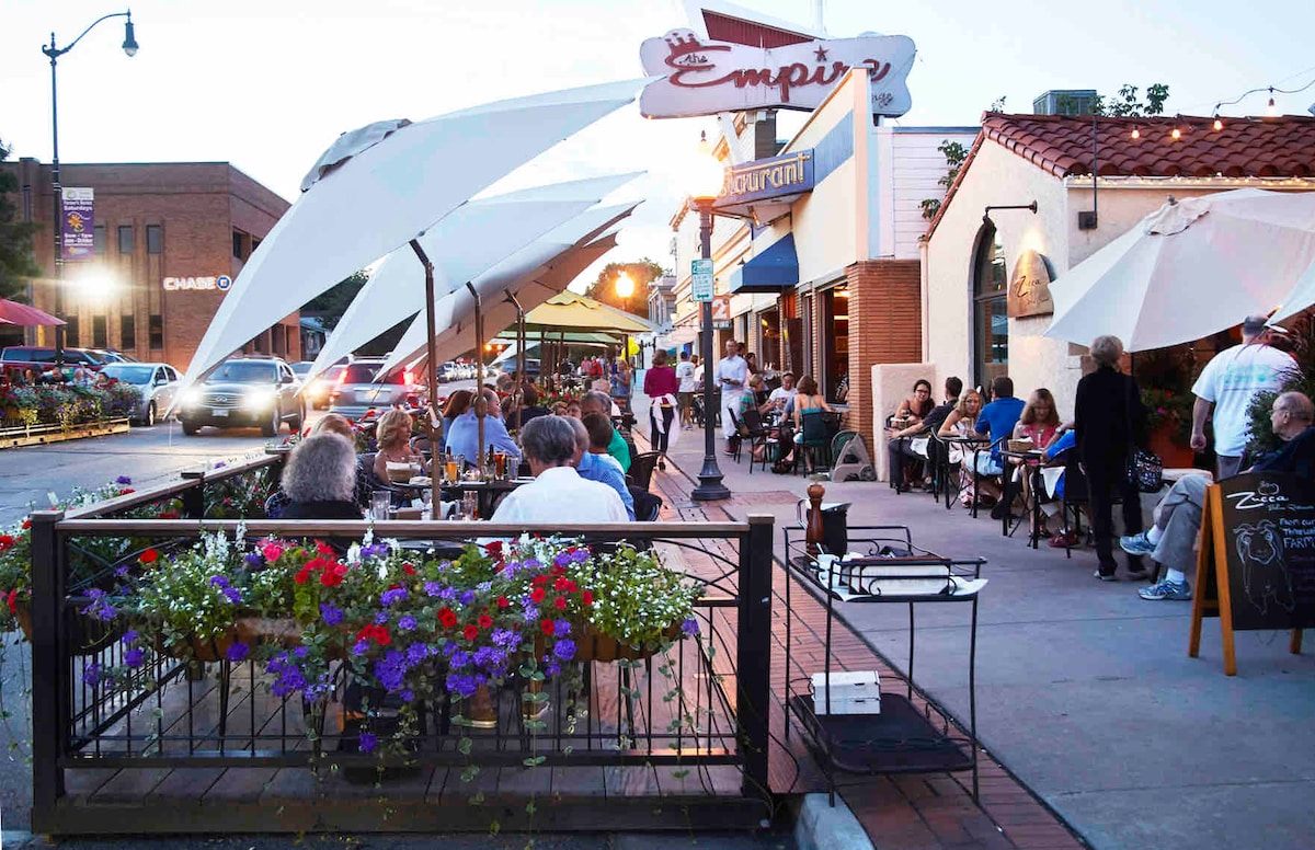 A lively street scene features a variety of outdoor dining options under large white umbrellas. Colorful flower pots adorn the patio area, while people enjoy meals at rustic wooden tables. Warm lighting reflects off nearby storefronts, contributing to a welcoming atmosphere.