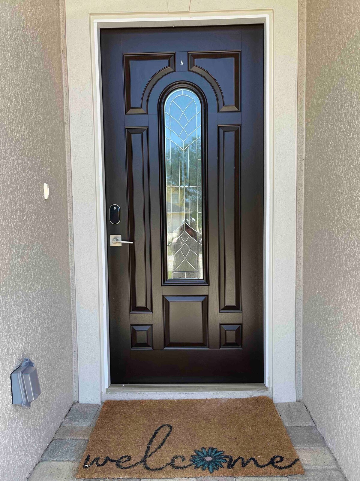A dark brown front door features an elegant arched window with decorative glass detailing. A welcome mat is positioned in front, displaying the word 'welcome' with floral accents. The entryway is surrounded by light-colored walls, providing a friendly first impression.