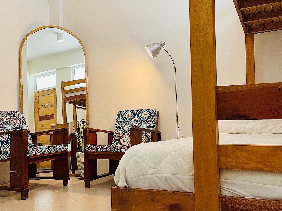 A cozy room is presented with wooden furniture, featuring a double deck bed and two upholstered chairs. Natural light floods the space from large windows, enhancing the earthy tones throughout. An arched mirror reflects the inviting setup, creating a sense of openness.
