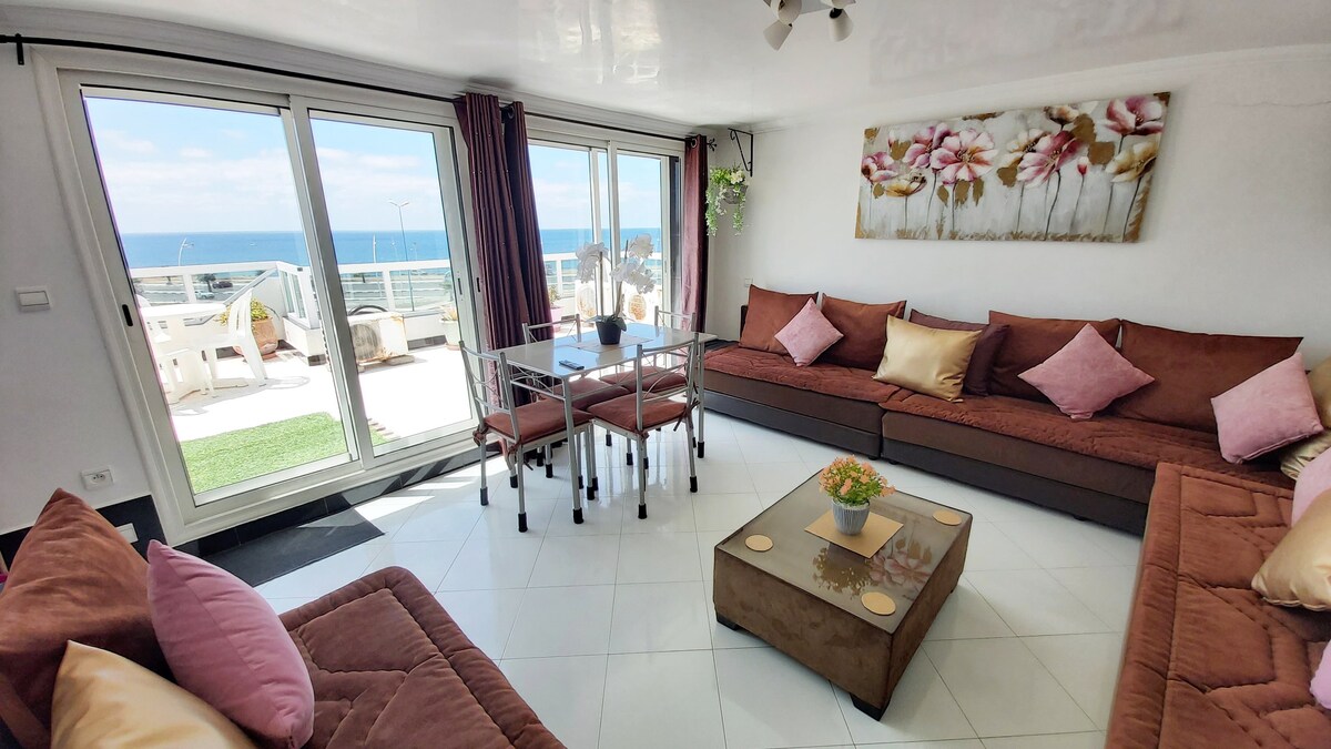 A light-filled living room showcases large glass doors that open to a sea view. Comfortable seating is arranged around a low coffee table, complemented by decorative cushions. A dining table is set for smaller gatherings, and floral artwork enhances the serene atmosphere.