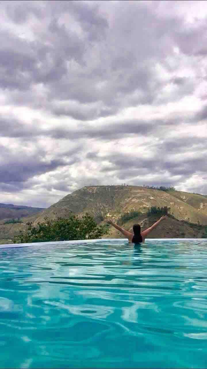 A serene infinity pool merges with the view of rolling hills under a cloudy sky. The tranquil water reflects the grey clouds above, creating a harmonious connection with the natural landscape. A person with arms raised enjoys the expansive scenery, emphasizing relaxation.