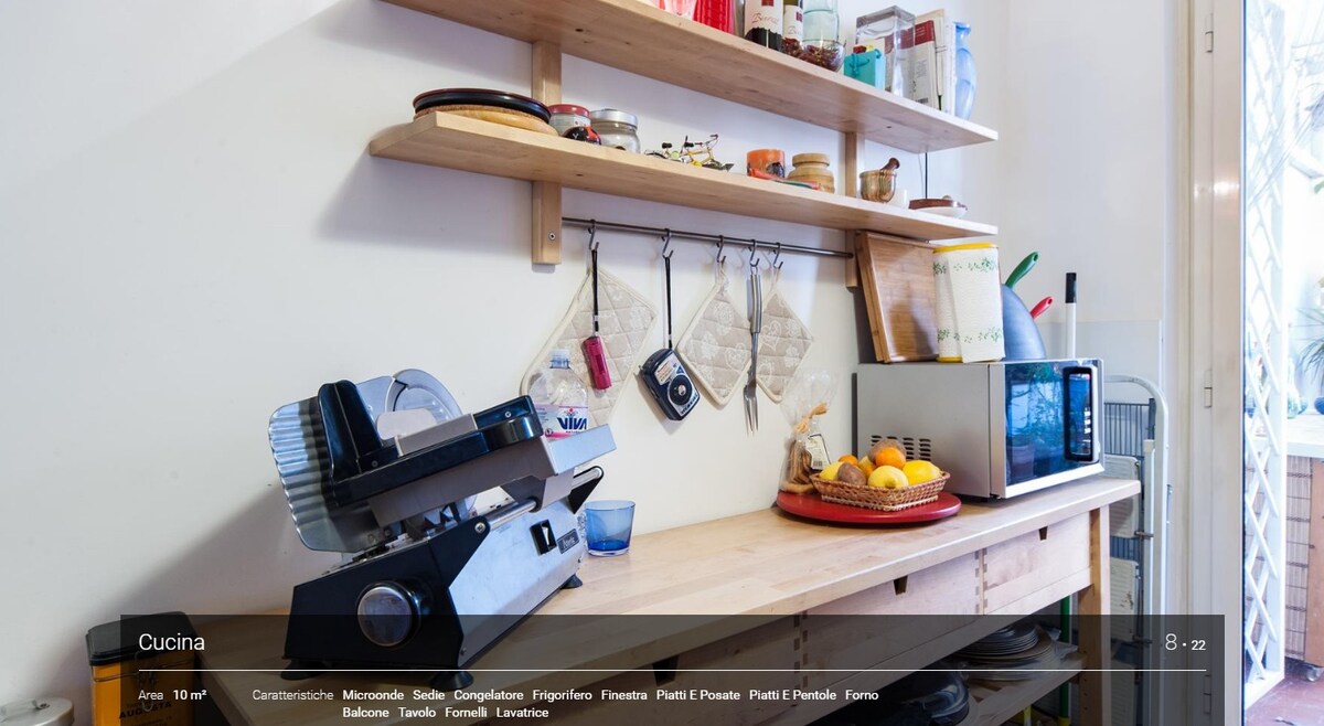 A functional kitchen space is displayed with wooden shelving holding kitchenware and decorative items. A small microwave and an orange juicer are visible on the countertop. A collection of fresh fruits adds a touch of colour to the area, creating an inviting cooking environment.