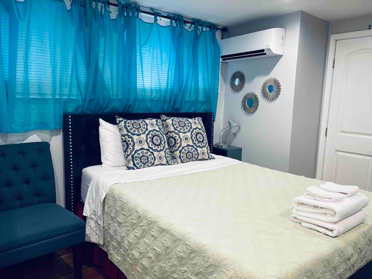 A well-lit sleeping area features a spacious bed with decorative pillows. A light blanket is neatly arranged at the foot of the bed. A blue curtain gently filters sunlight. A small chair and side table complement the space, while a modern air conditioning unit is mounted on the wall.