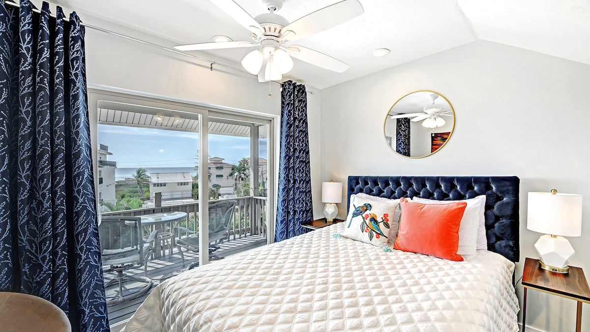 A bright loft bedroom features a spacious bed with a patterned duvet and colorful pillows. Large sliding doors provide access to a balcony with views of the Gulf. Two bedside lamps and decorative curtains complete the inviting space.