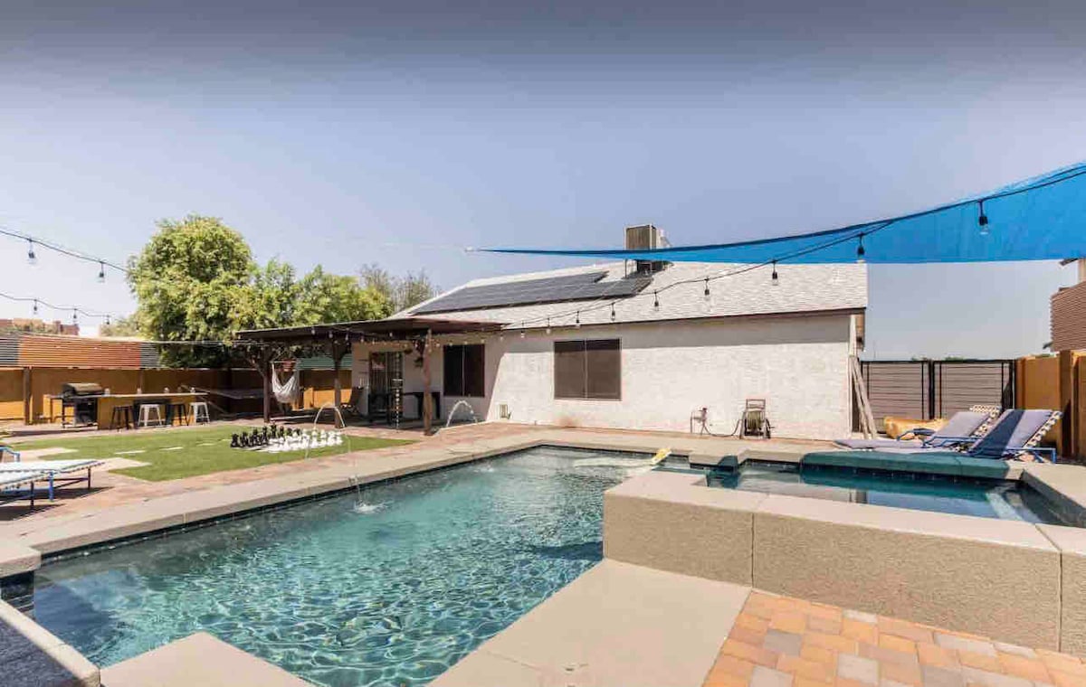 A spacious backyard features a refreshing swimming pool and a separate spa, surrounded by decorative pavers. Comfortable lounge chairs are situated by the pool, and a shaded area is provided by a blue fabric canopy. Lush greenery adds a natural touch to the outdoor space.