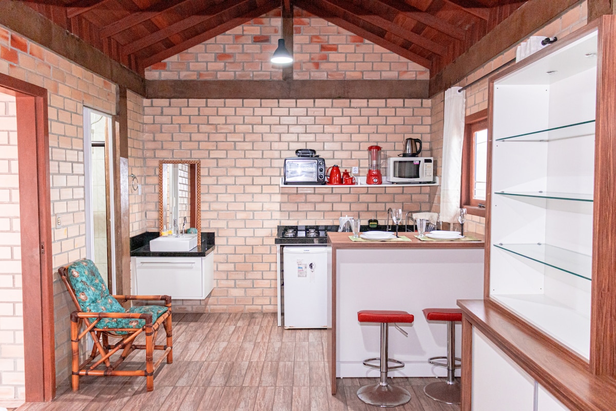 The kitchen area features modern appliances including a stove, microwave, and refrigerator, set against a backdrop of rustic brick walls. A dining space with bar stools is positioned beside a countertop. A comfortable chair completes the inviting interior layout.