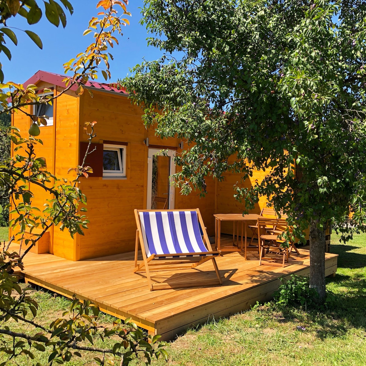 A wooden deck is featured outside a brightly colored tiny house, with a striped lounge chair positioned for relaxation. A small table and chairs are arranged nearby under the shade of a tree, creating a serene outdoor space for enjoyment.