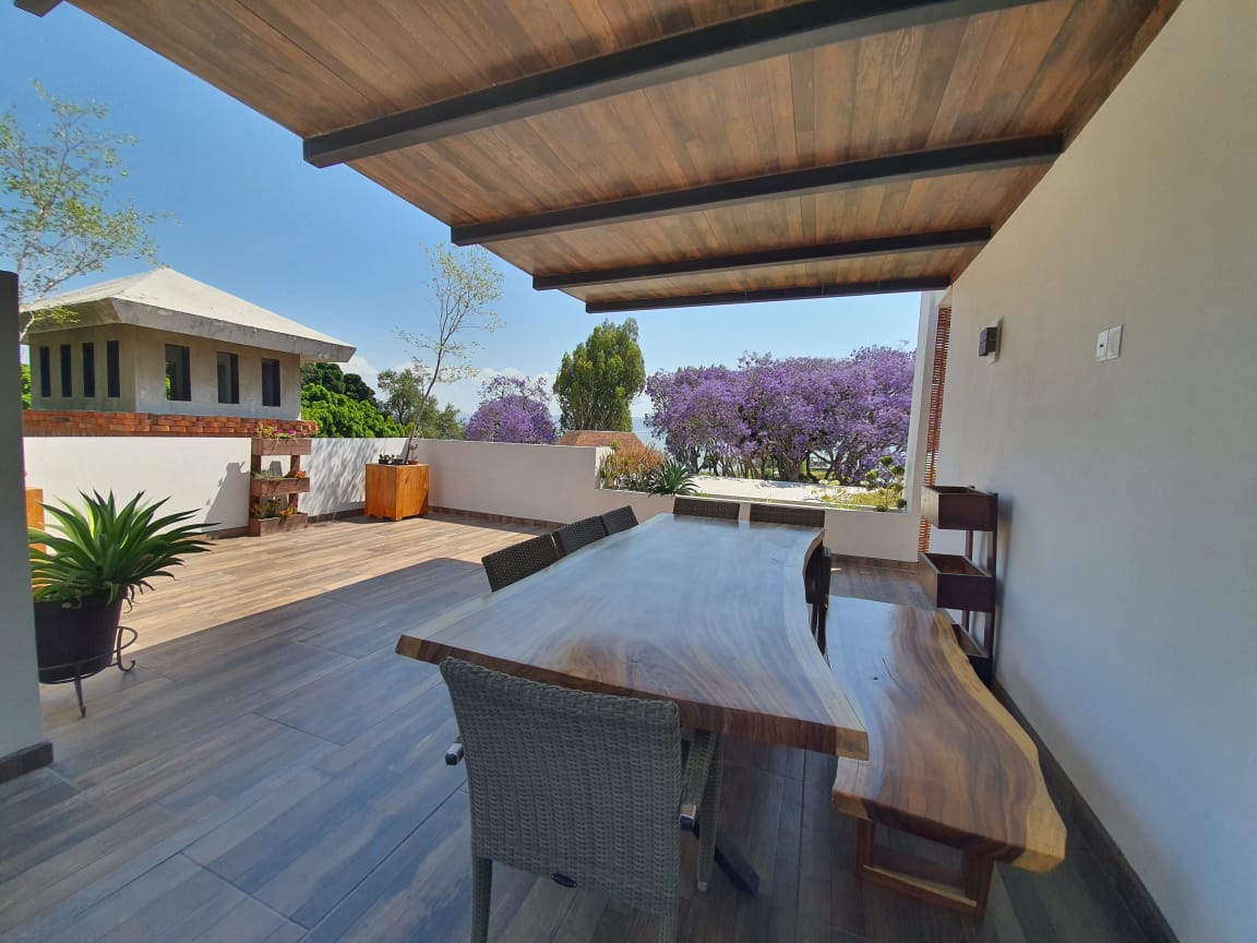 An outdoor dining area features a large wooden table surrounded by woven chairs, placed under a wooden pergola. Lush greenery and blooming jacaranda trees are visible in the background, contributing to the serene atmosphere.