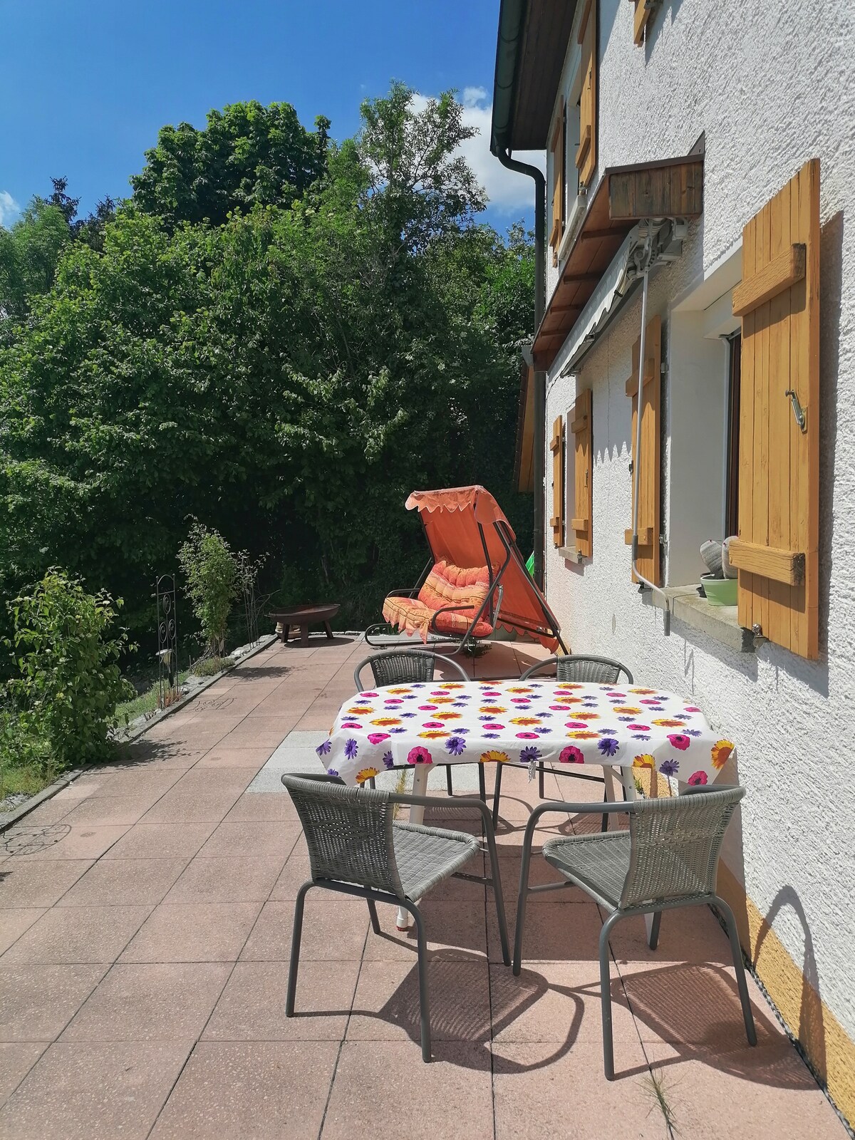 A terrace features a table with colorful tablecloth and four wicker chairs. Nearby, a cushioned orange swing is positioned under a bright sky, surrounded by greenery. Thick trees provide privacy and contribute to the serene outdoor setting.