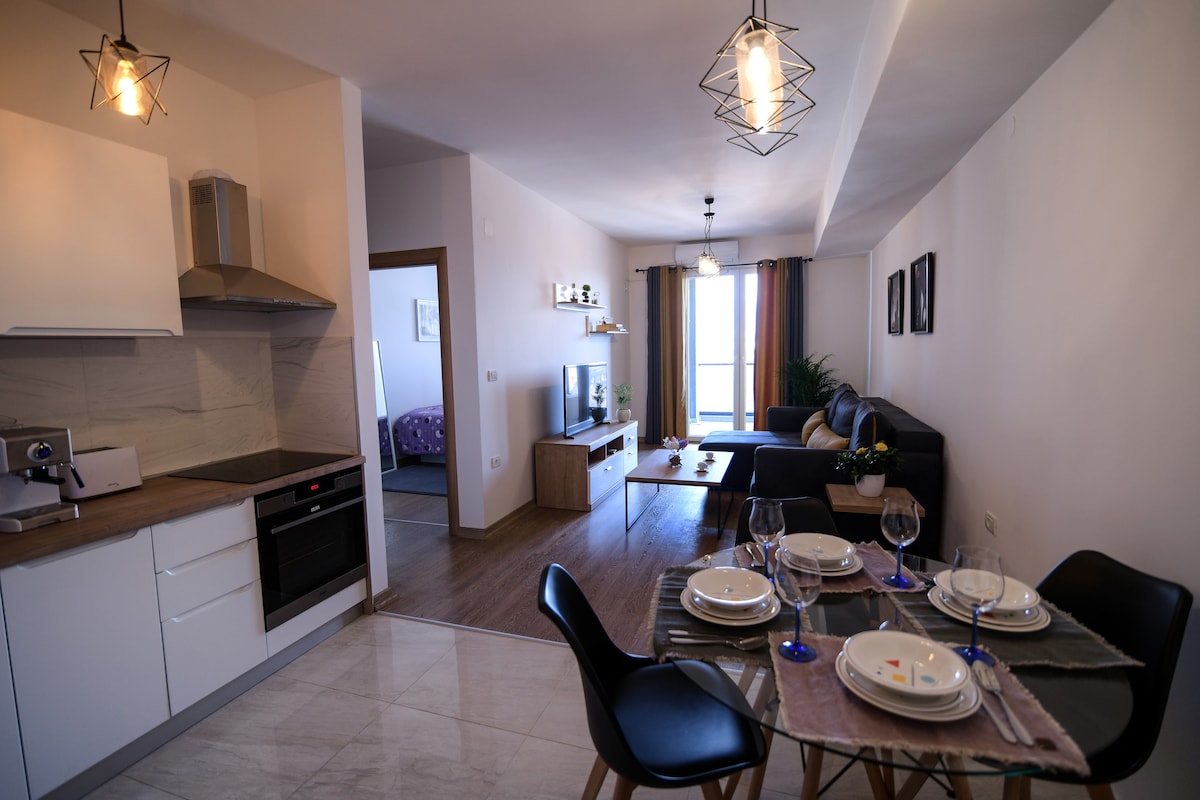 A modern apartment interior is shown, featuring a dining area with a table set for four. The kitchen is equipped with a stovetop and oven, while a comfortable sofa is placed in a cozy lounge area that receives natural light from a large window.