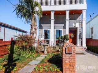 The exterior of a contemporary home showcases a welcoming entrance with a brick accent and a mailbox. Pathways lined with stone lead to the entrance, and seasonal plants are arranged around a decorative fountain. Tall palm trees add greenery to the surroundings.
