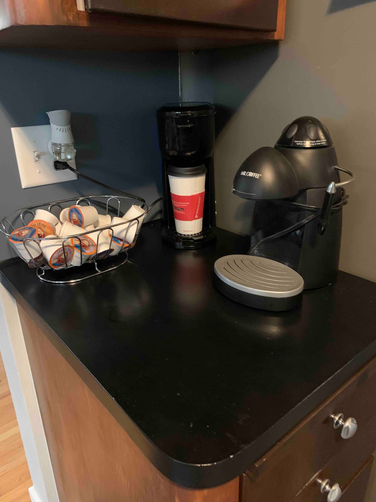 A kitchen countertop is equipped with a coffee maker and an espresso machine, both in black. A basket containing assorted coffee pods is displayed alongside a cup. The surface is dark and smooth, providing a clean and organized appearance.