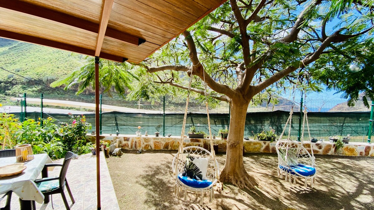 An outdoor area is highlighted by two suspended swings with blue cushions, nestled beneath a large tree. A table with chairs is positioned nearby, surrounded by greenery. The backdrop features a landscape of hills and a protective fence, enhancing the tranquil setting.