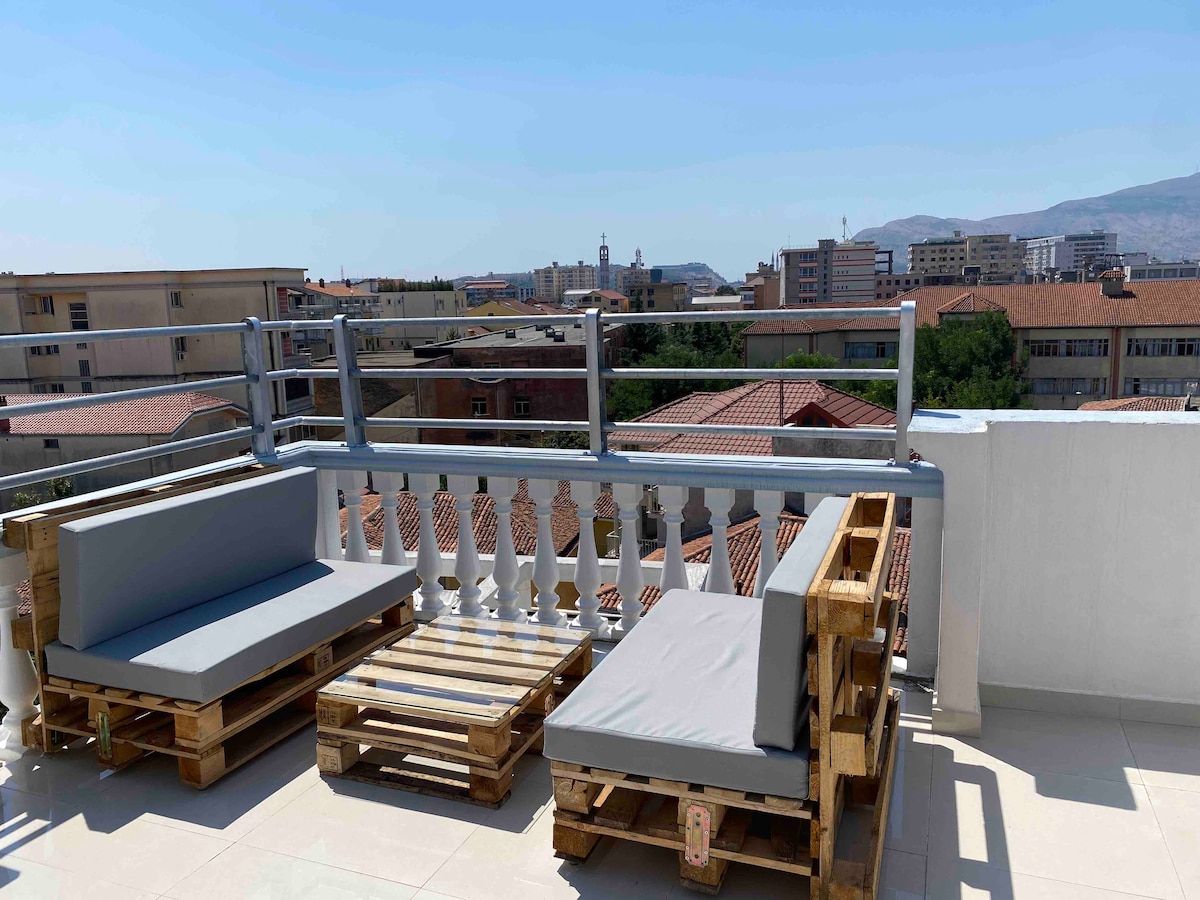 An outdoor rooftop deck features comfortable seating made from wooden pallets and cushions. A white balcony railing offers a view of the surrounding rooftops and distant buildings, under a clear blue sky.