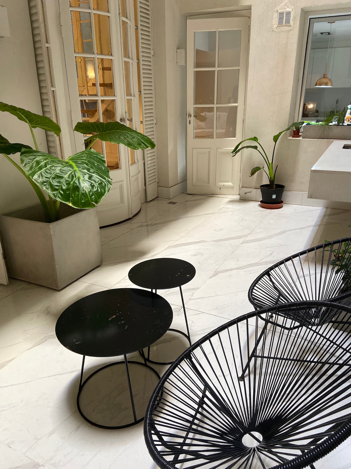 A serene patio is presented with smooth marble flooring, complemented by large potted plants. Two black accent tables are visible alongside a pair of black woven chairs. French doors allow for natural light, enhancing the overall brightness of the space.
