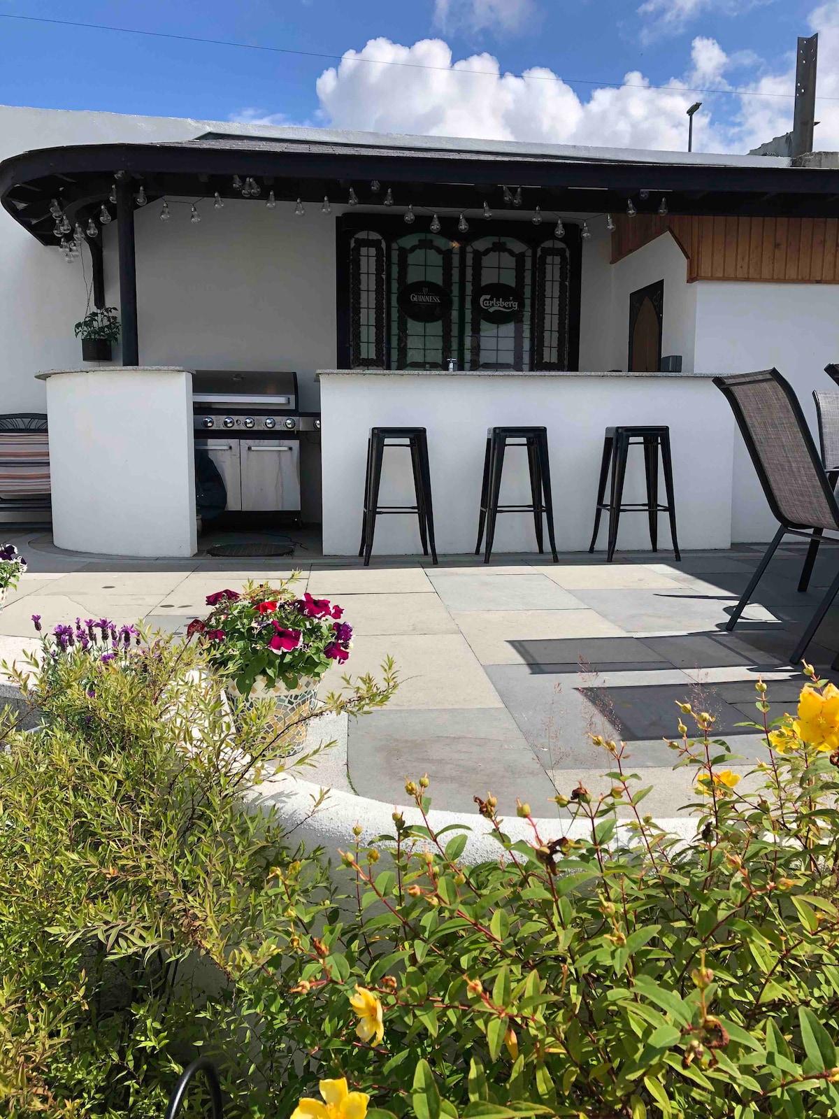 A spacious outdoor area features a sleek barbecue setup beneath an overhang. High-top stools surround a bar-like counter, with colorful flowers in the foreground adding a vibrant touch. A sunny blue sky with scattered clouds provides a bright backdrop.