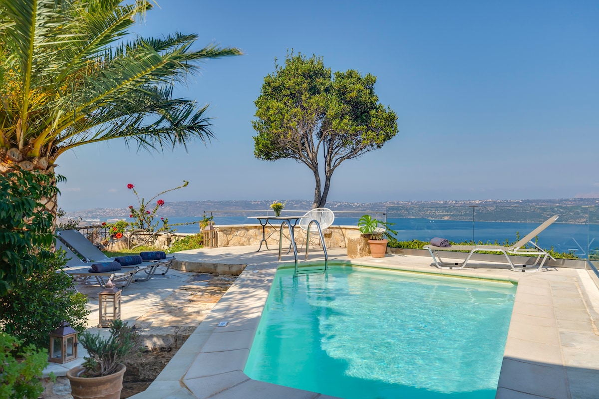 An inviting outdoor pool area features a clear blue pool surrounded by sunbeds and lush greenery. A tree provides shade, while potted plants add a touch of color. The panoramic view overlooks the serene waters of Souda Bay, enhancing the tranquil environment.