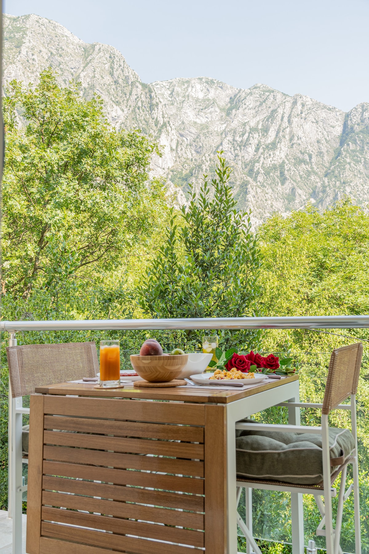 A dining table with wooden accents is positioned on a balcony, offering views of surrounding lush greenery and mountains in the background. The table is set with a bowl of fruit, a plate of salad, and a glass of orange juice.