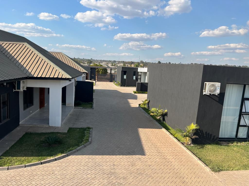The image shows a view of a modern courtyard with multiple buildings featuring varied architectural styles. A paved pathway runs through the center, flanked by patches of green grass and low-growing plants. Clear blue skies and fluffy clouds complete the serene setting.