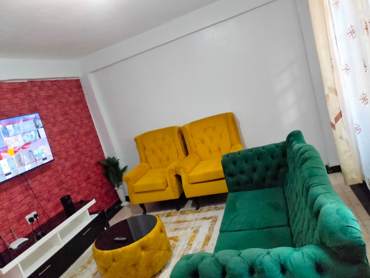 The living area showcases a green sectional sofa paired with yellow accent chairs, creating a striking color contrast. A round coffee table rests in the center on a patterned rug. A wall-mounted television is visible, while light filters through sheer curtains.