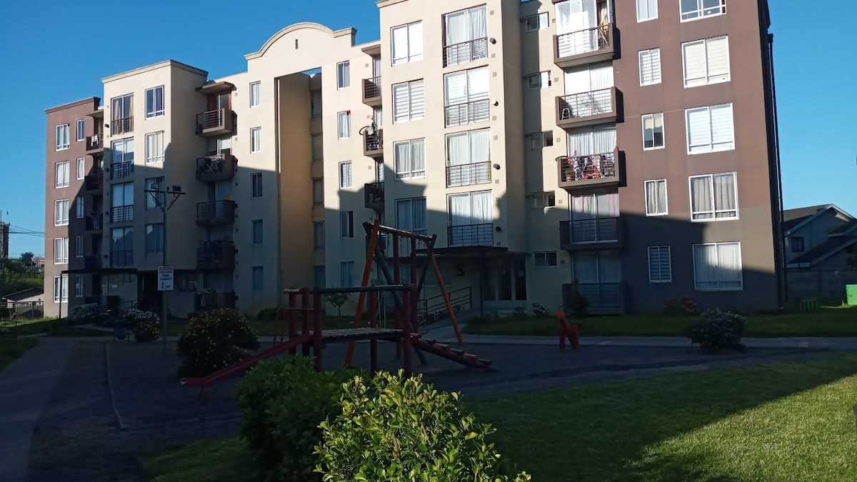 A multi-story apartment building is viewed from the outside, featuring a well-maintained courtyard. Play equipment is visible in the foreground, surrounded by landscaped green areas and flowering shrubs, with balconies on the upper levels extending outward.